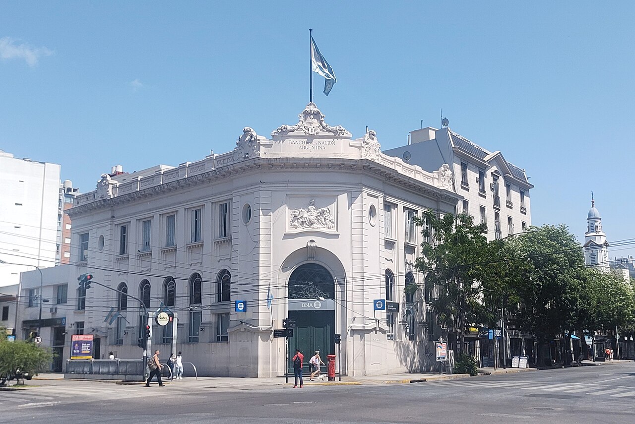 White corner building of a Banco de la Nación Argentina branch with a BNA sign over the door and an Argentine flag on the roof