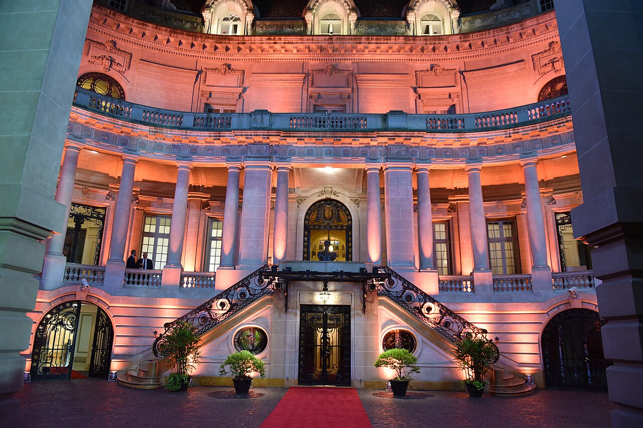 The floodlit inner courtyard of the Palacio San Martín in Buenos Aires, with a curved stone façade, columns, a double staircase and a red carpet