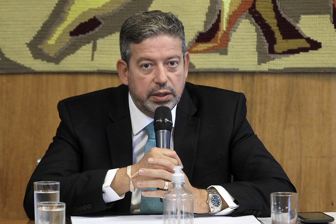 Arthur Lira in a dark suit and teal tie speaks into a microphone at a table, in front of a woven wall hanging
