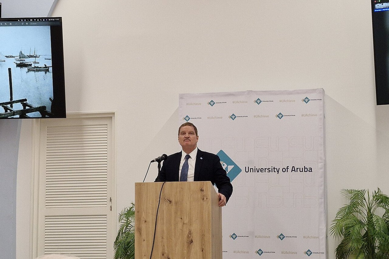 Mike Eman in a dark suit and blue tie stands at a wooden lectern before a University of Aruba banner, with old harbour photos on a screen