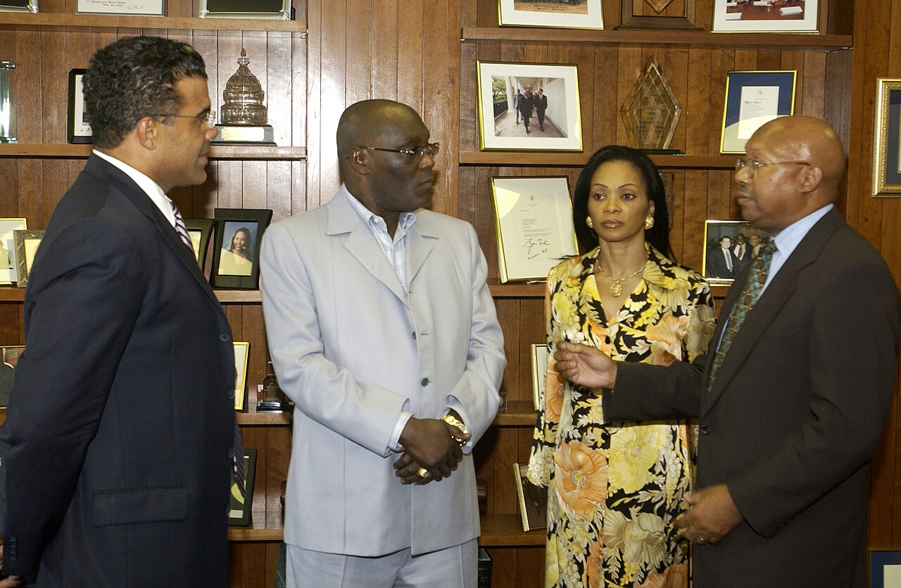 Atiku Abubakar, in a light grey suit, stands in a wood-panelled office with three other people, two men in dark suits and a woman in a floral dress, during a 2003 meeting