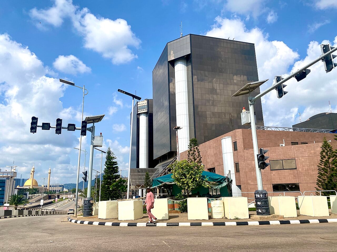 The dark glass NNPC Towers in Abuja with white cylindrical columns, traffic lights in front and the golden dome of the National Mosque in the distance