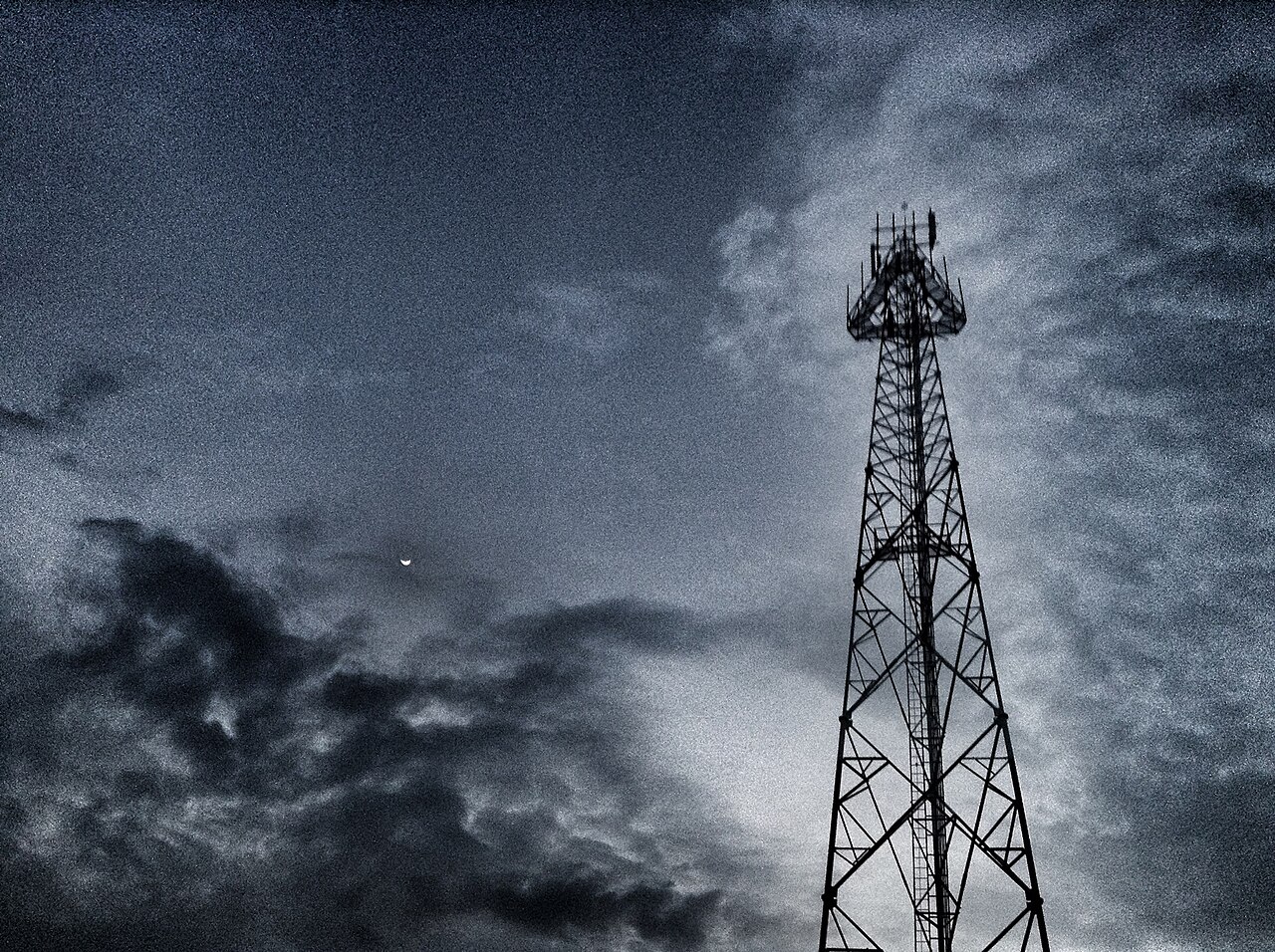 A steel lattice mobile phone antenna tower silhouetted against a cloudy evening sky with a crescent moon
