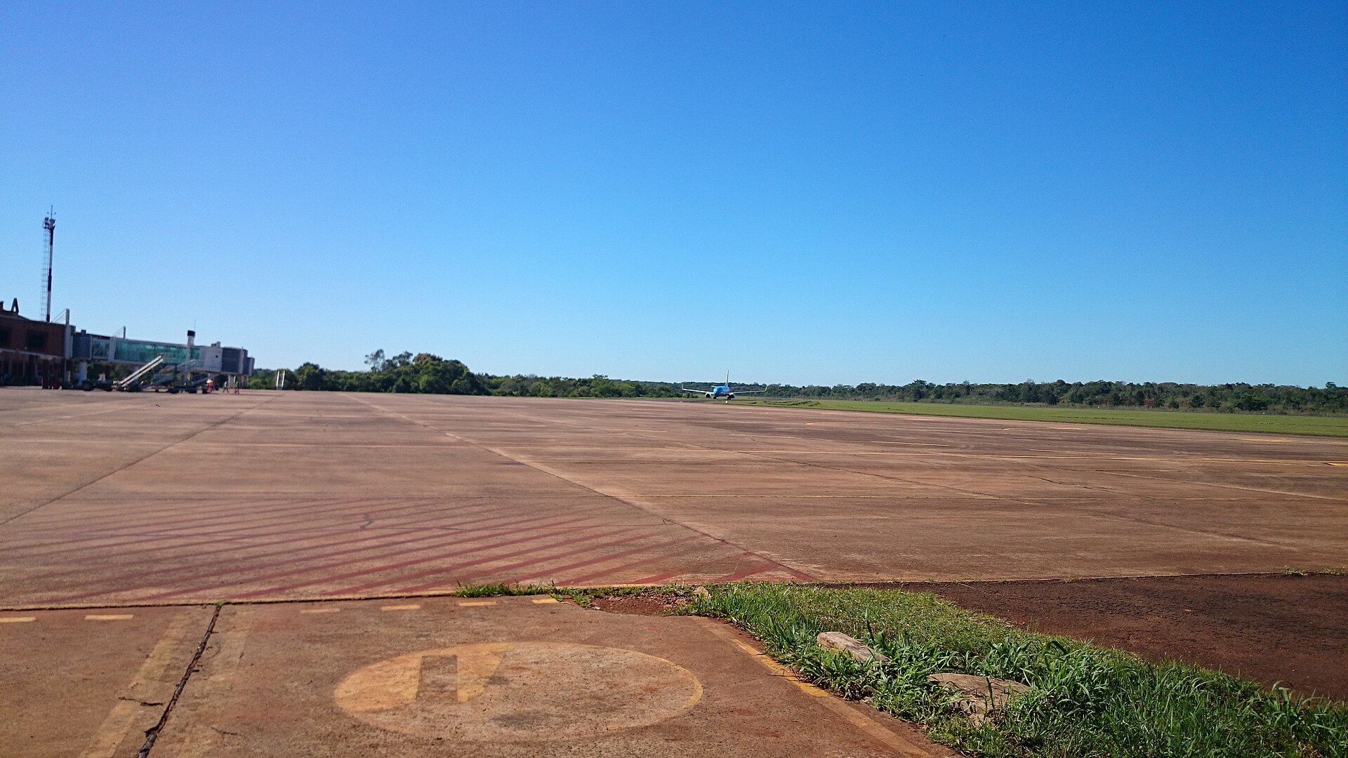 Wide apron and runway of Cataratas del Iguazu International Airport with a small terminal and a distant airliner under a blue sky