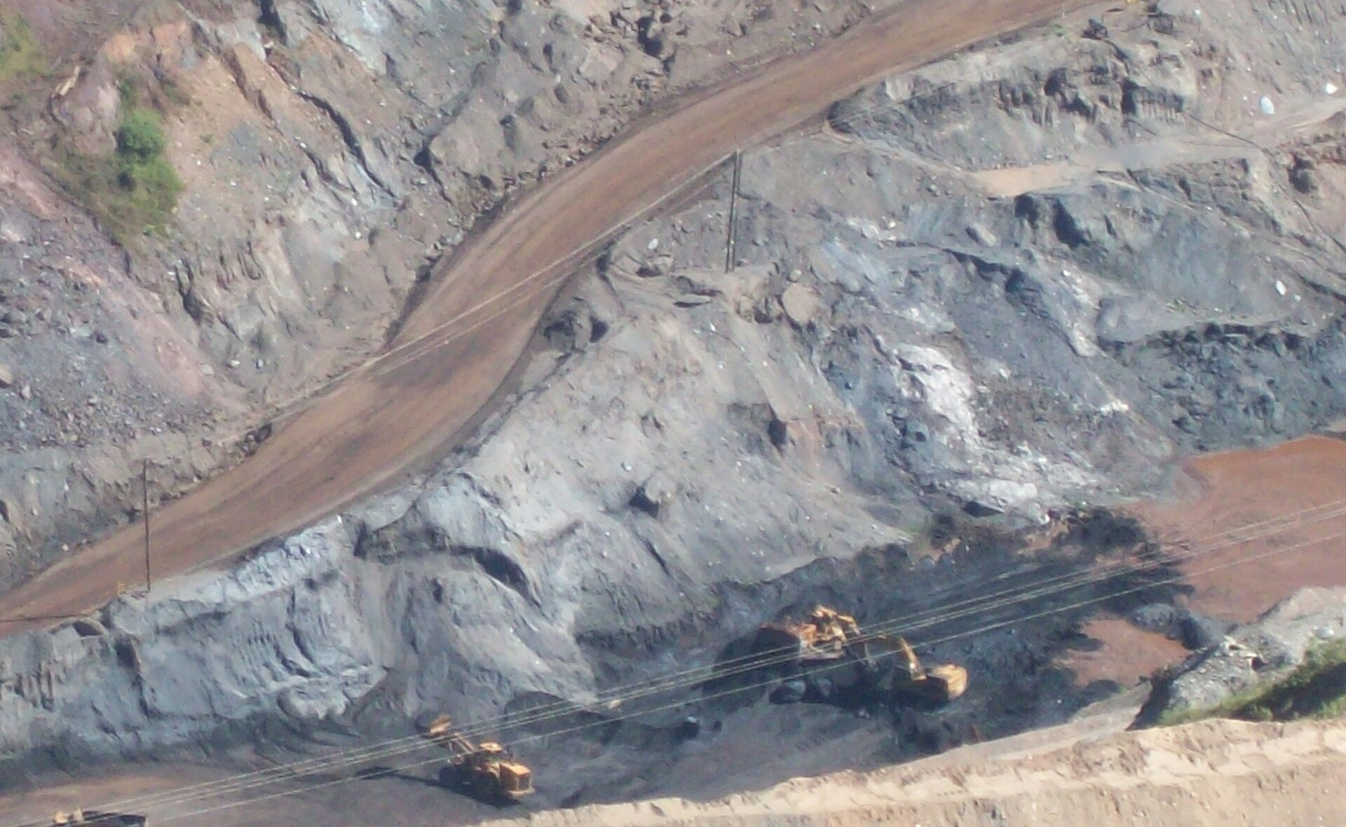 Yellow excavators and trucks work on terraced grey and red rock walls of an open-pit mine near Itabira