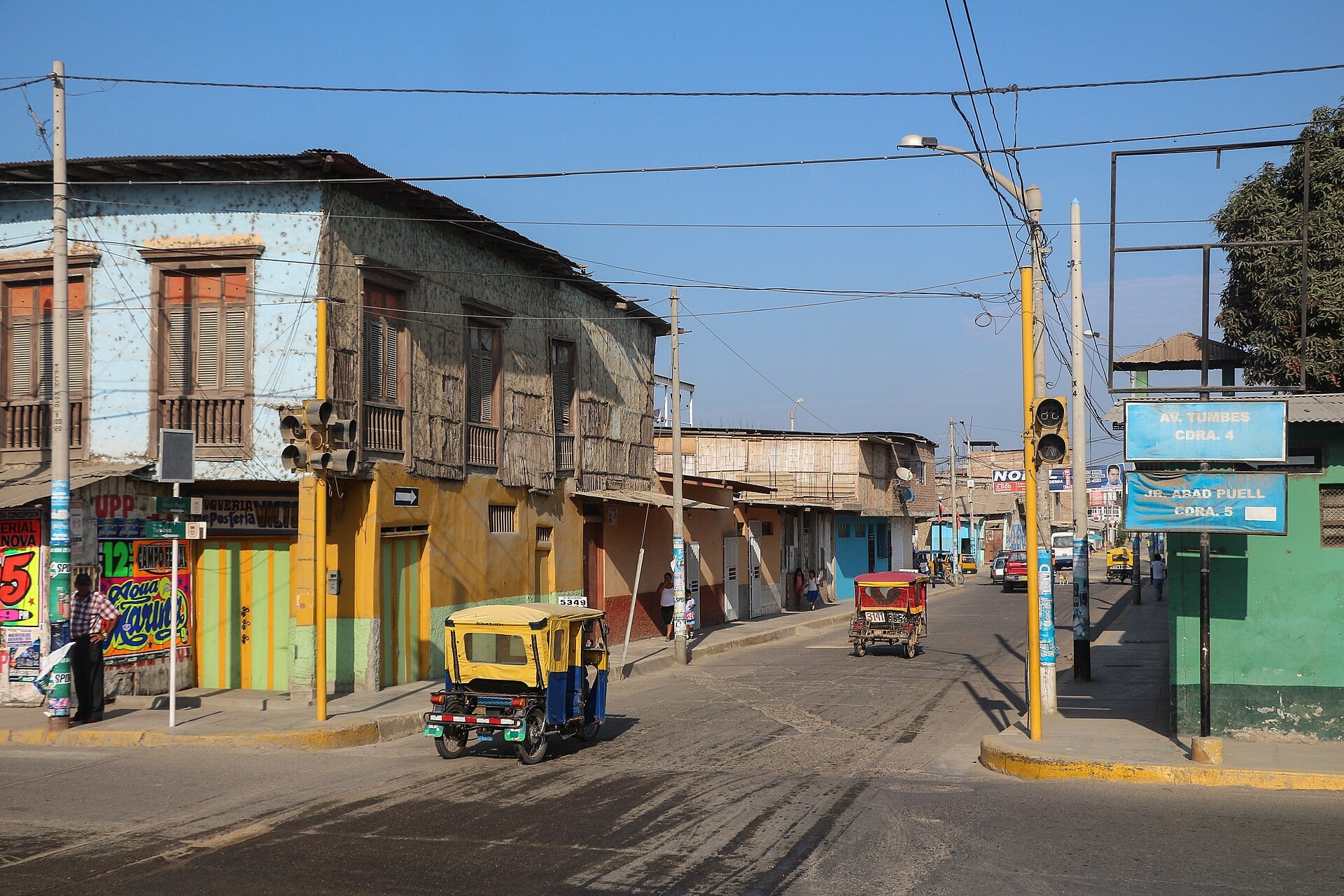 A yellow motorcycle taxi turns at a street corner lined with weathered wooden-balcony houses in Tumbes, Peru