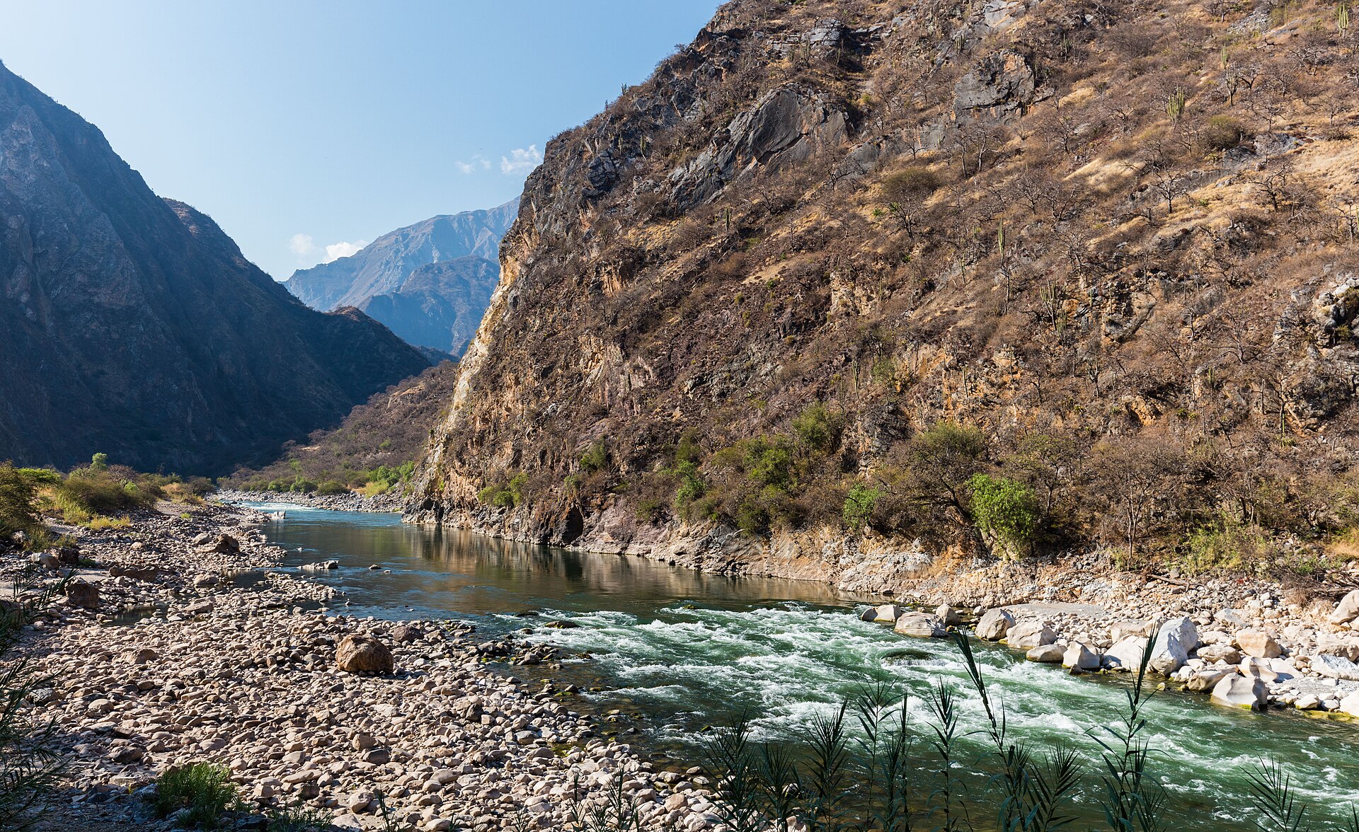 The Apurimac river runs green and white over rapids between steep, dry mountain slopes with a rocky bank in the foreground