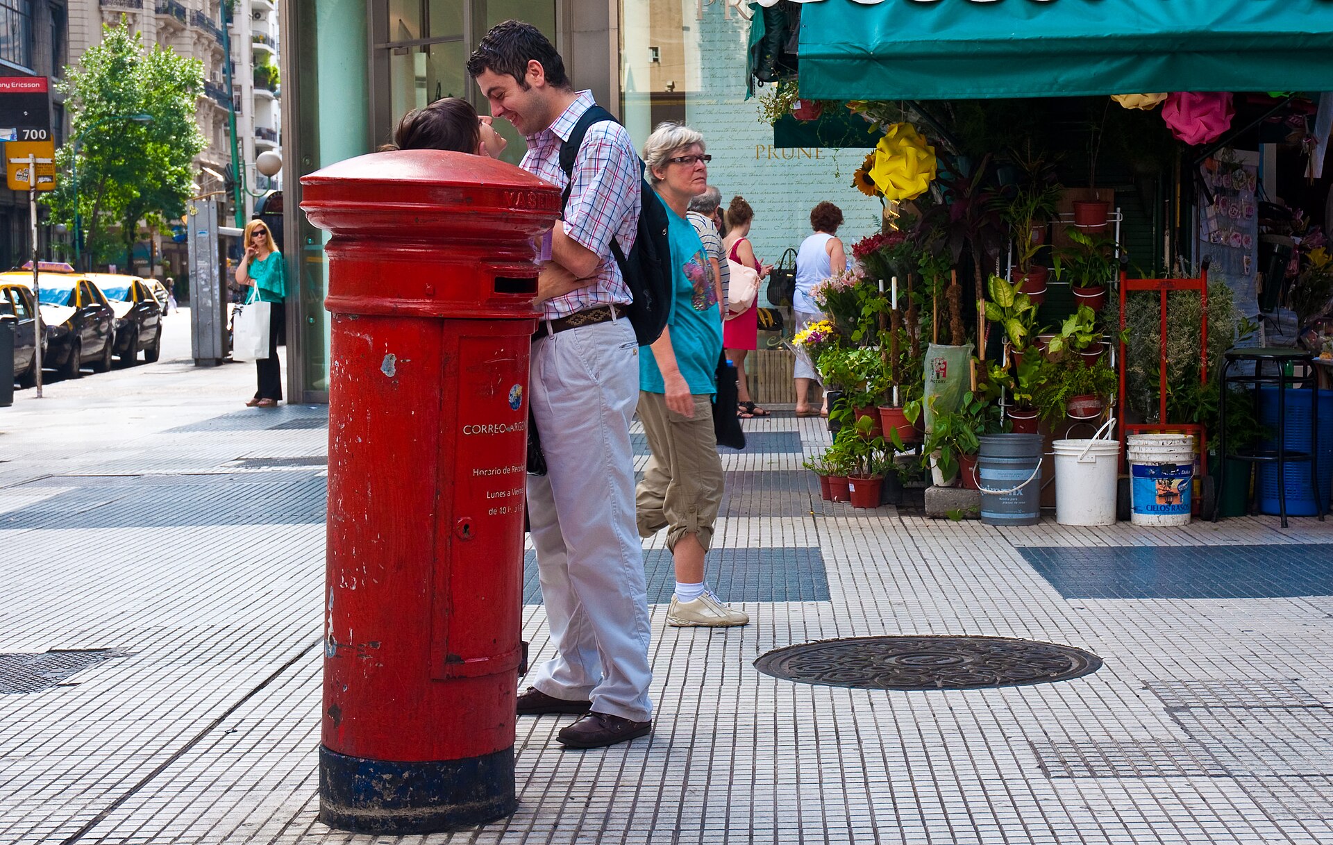 A red postbox and pedestrians on a paved downtown corner on Avenida Córdoba in Buenos Aires, with a flower stall at right