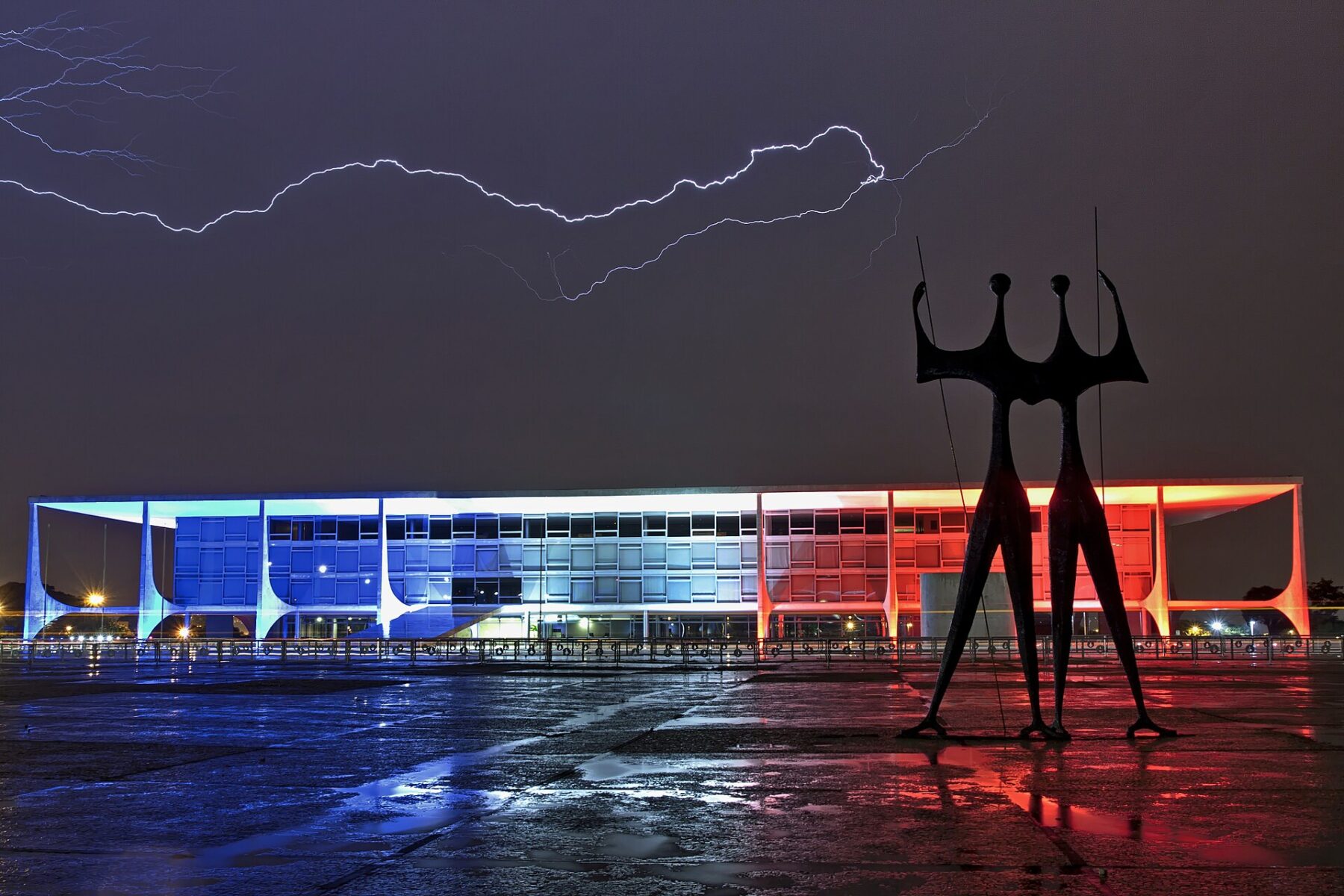 The Planalto Palace in Brasilia lit in blue and red at night under a lightning bolt, with the Warriors sculpture in front