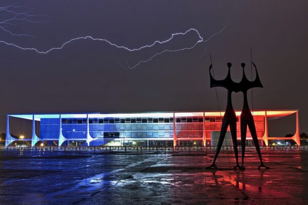 The Planalto Palace in Brasilia lit in blue and red at night under a lightning bolt, with the Warriors sculpture in front