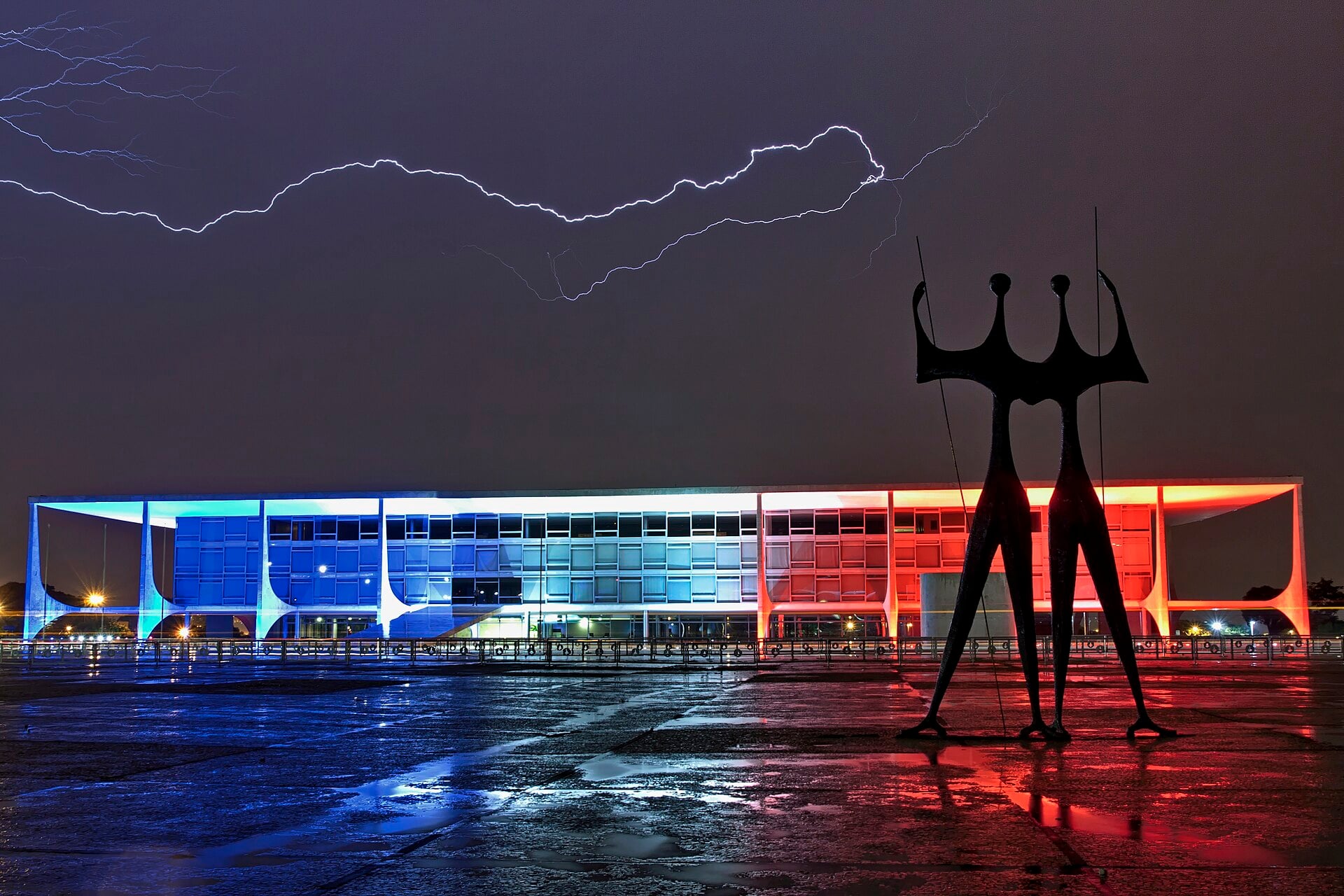 The Planalto Palace in Brasilia lit in blue and red at night under a lightning bolt, with the Warriors sculpture in front
