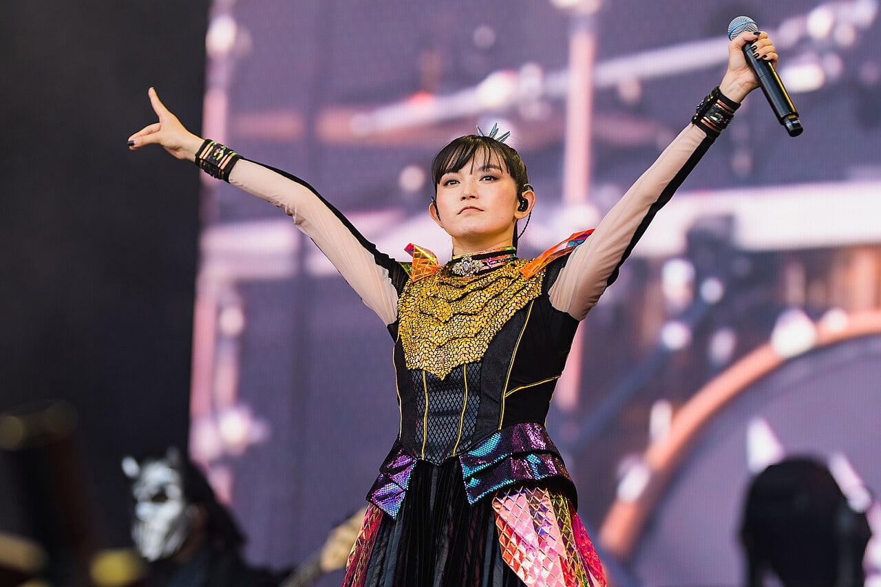 A Babymetal singer with arms raised and a microphone in one hand, in a black and gold stage costume, on stage at a festival.