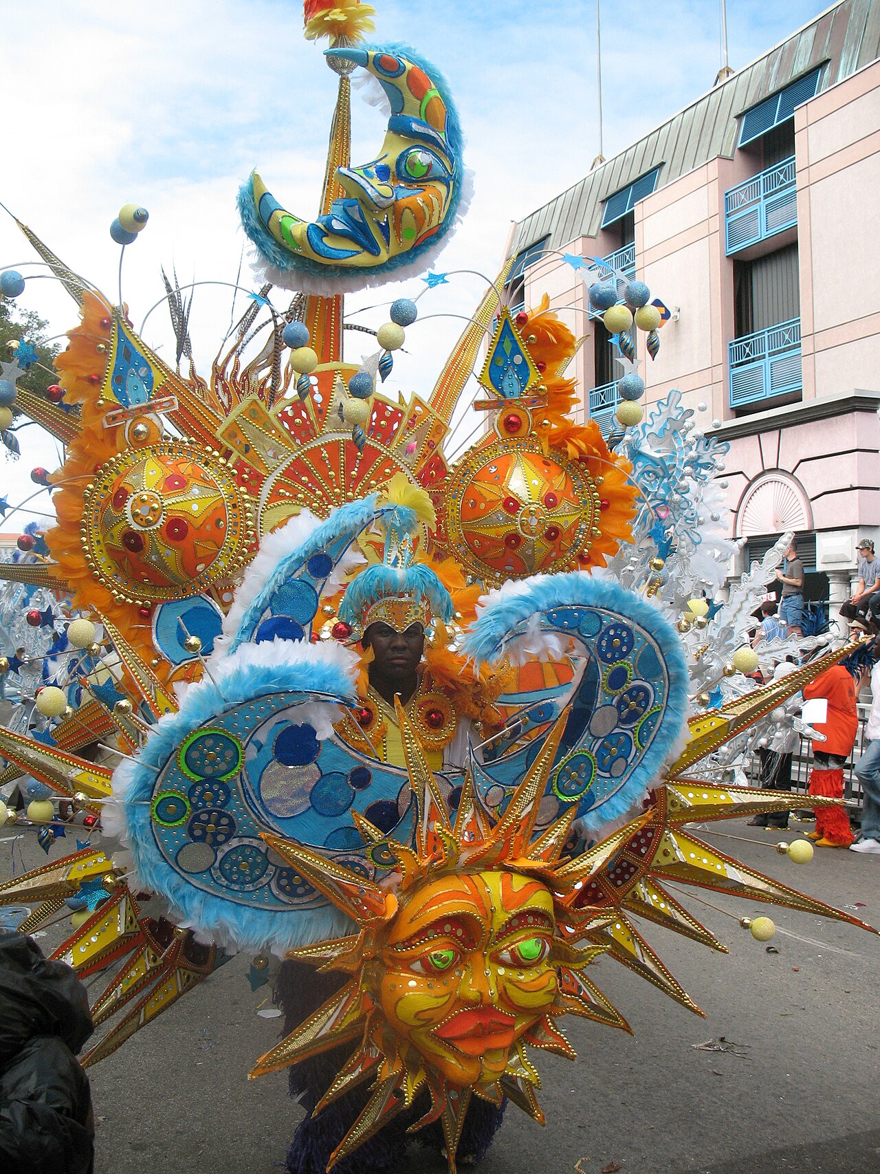 Dancer inside a towering Junkanoo costume of orange sun and blue moon faces with feathers on a street