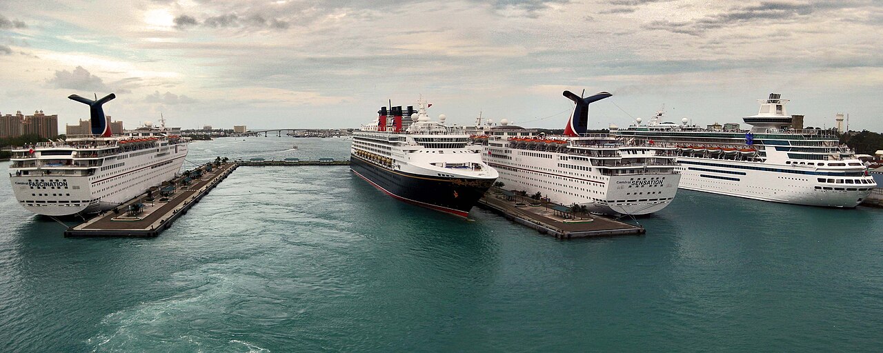 Four cruise ships moored side by side at Prince George Wharf in Nassau harbour under a cloudy sky, with a bridge and hotel towers in the distance.