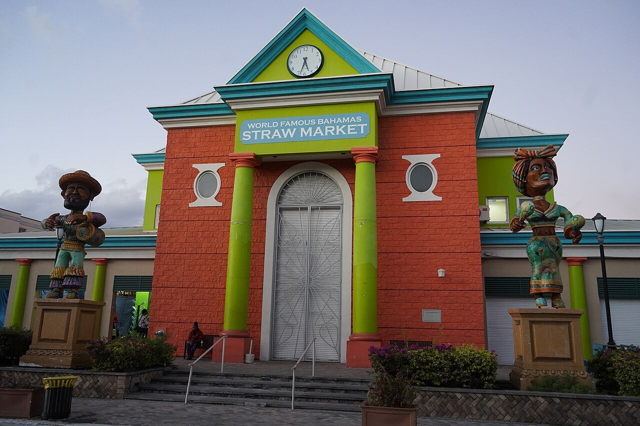 The colourful front of the Straw Market in downtown Nassau, with a green and orange facade, a clock, a sign reading World Famous Bahamas Straw Market and two painted statues.