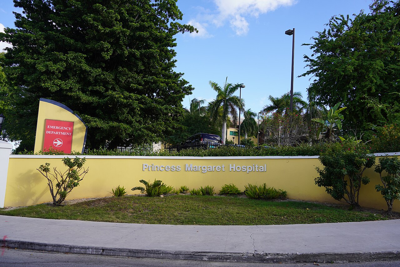 The Princess Margaret Hospital sign on a yellow wall beside a red Emergency Department sign, with palm trees and a blue sky behind, in Nassau