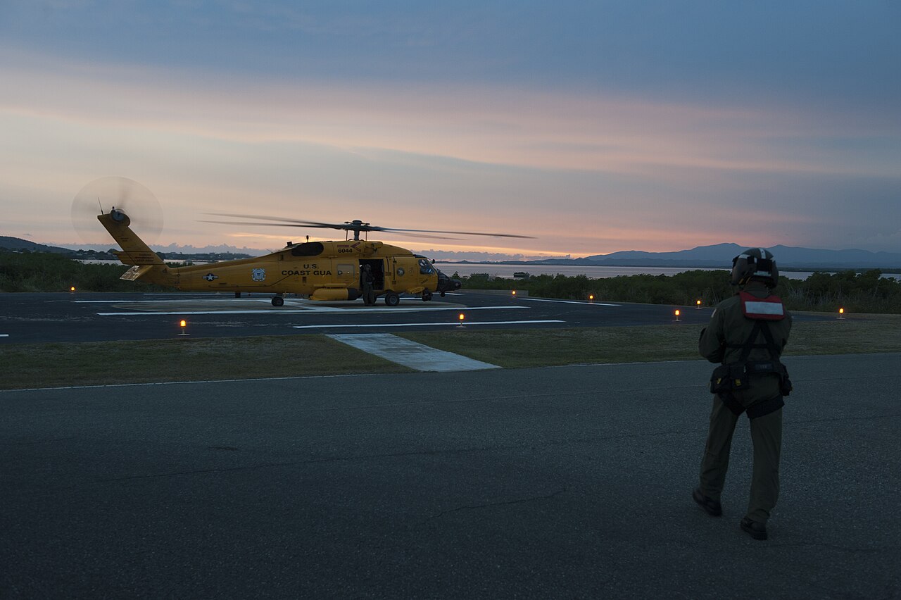 A US Coast Guard MH-60 Jayhawk helicopter parked on a helipad at dusk in the Bahamas, with a crew member in a flight helmet walking toward it