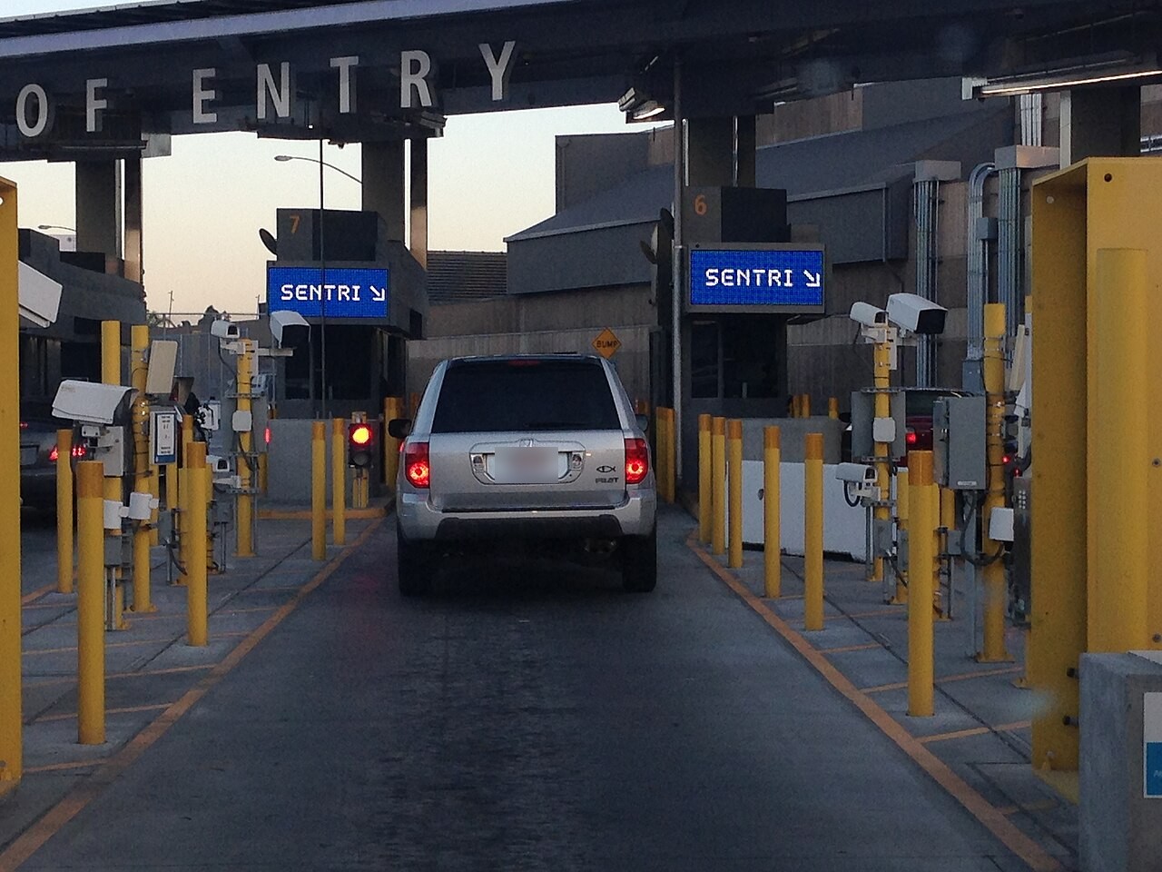 A silver SUV waits in a lane under the Port of Entry canopy at San Ysidro, with blue SENTRI signs, yellow posts and cameras