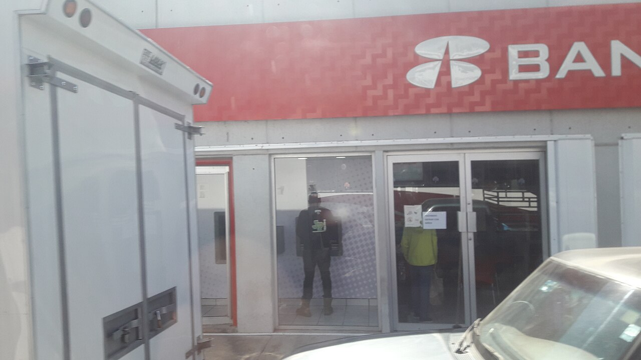 Red Banorte bank sign above the glass entrance of a branch, with a delivery van and a car in the foreground