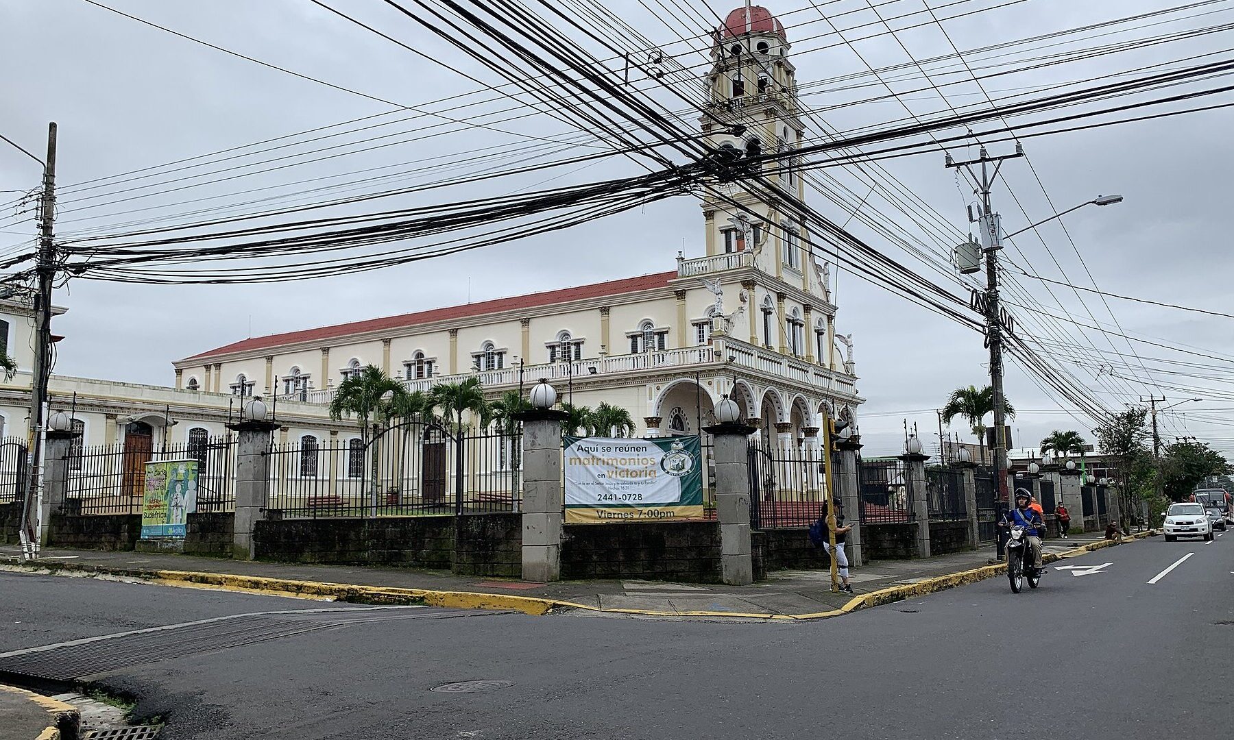 Cream-coloured church with a red roof and a tall bell tower on a street corner in Alajuela, Costa Rica, under an overcast sky