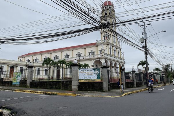 Cream-coloured church with a red roof and a tall bell tower on a street corner in Alajuela, Costa Rica, under an overcast sky