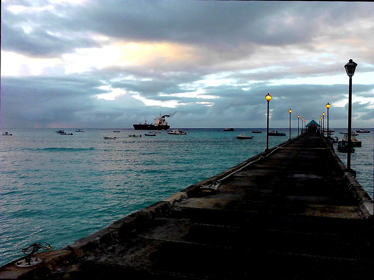 A pier with lit lamps at dawn over turquoise water and fishing boats at Oistins on the south coast of Barbados