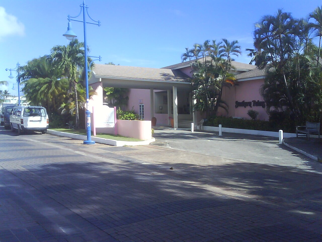Pink entrance building of the Southern Palms Beach Club among palm trees on a paved street in Saint Lawrence Gap, a tourist resort area on the south coast.