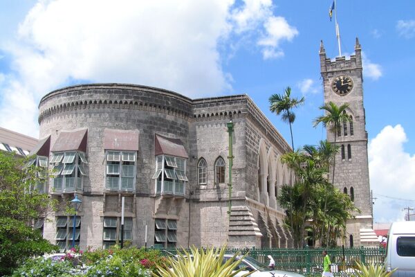 The Parliament Buildings in Bridgetown, a grey stone building with a round corner, shuttered windows and a clock tower with a flag, under a blue sky