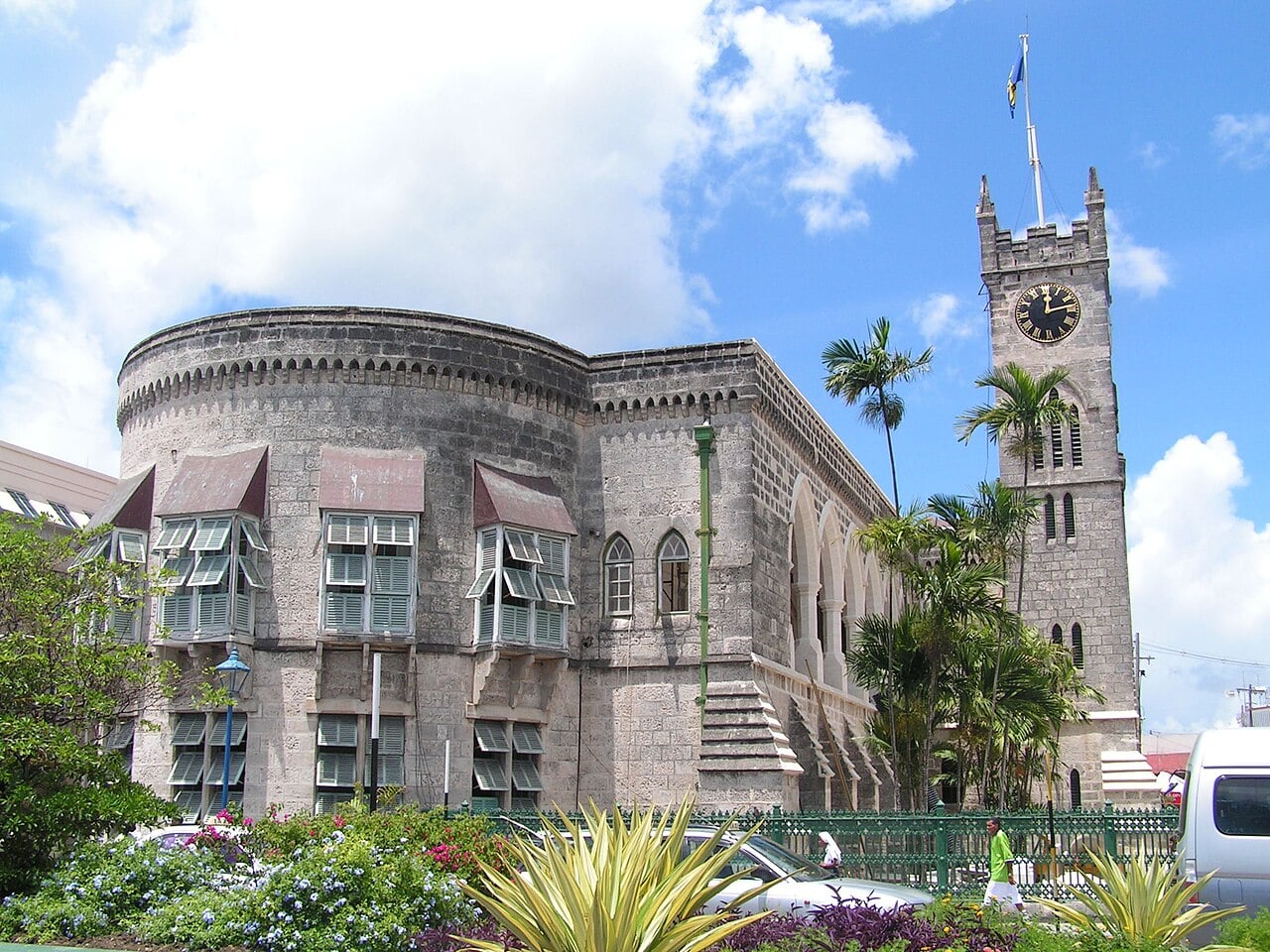 The Parliament Buildings in Bridgetown, a grey stone building with a round corner, shuttered windows and a clock tower with a flag, under a blue sky