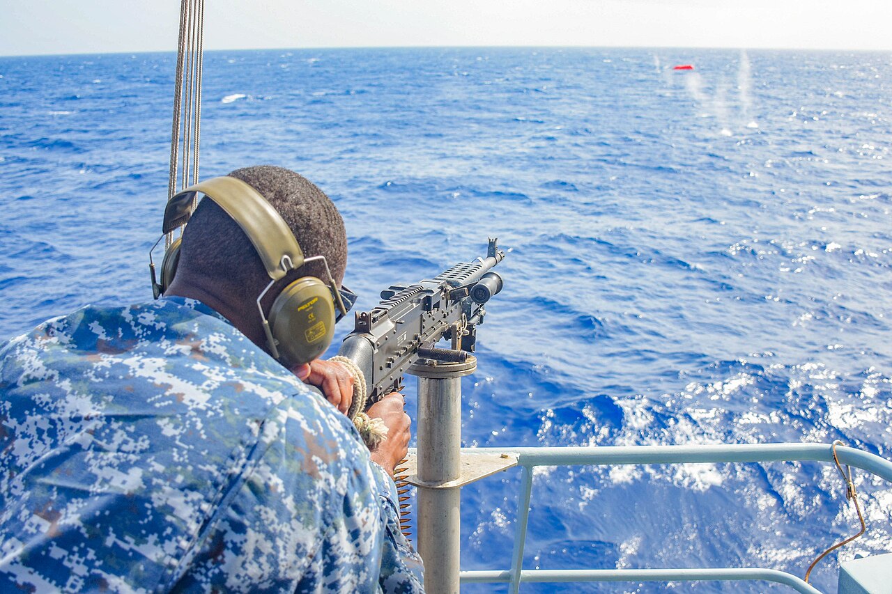 A Barbados Coast Guard sailor in blue camouflage and ear defenders fires a mounted machine gun at a target at sea