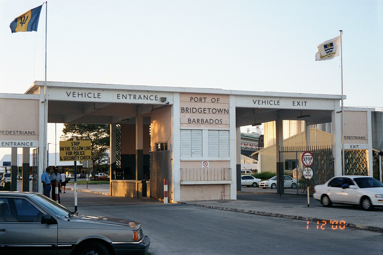 The gatehouse of the Deep Water Harbour at the Port of Bridgetown with vehicle entrance and exit gates, the Barbados flag and a cruise ship behind