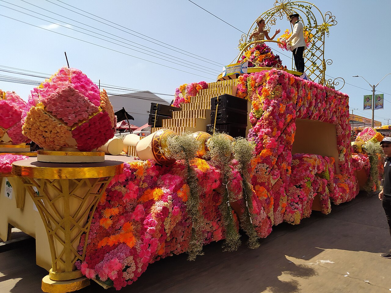 A float covered in pink, orange and yellow flowers with a queen and an escort in white on top, in a Carnival parade in Barranquilla.