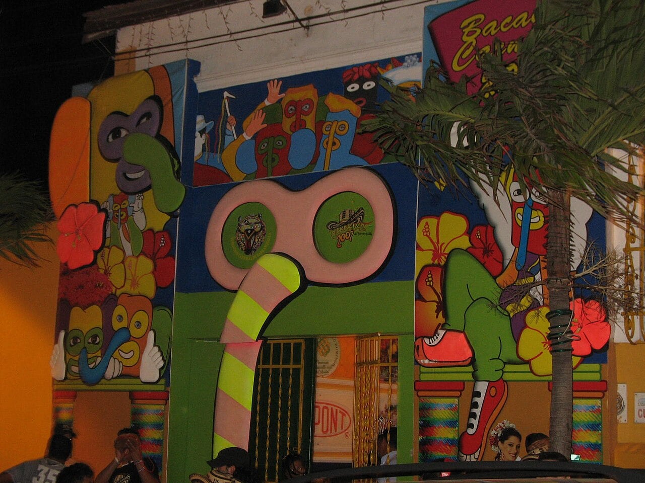 The painted front of the Casa del Carnaval in Barranquilla at night, with a giant Marimonda mask, flowers and a large green figure in red sneakers.