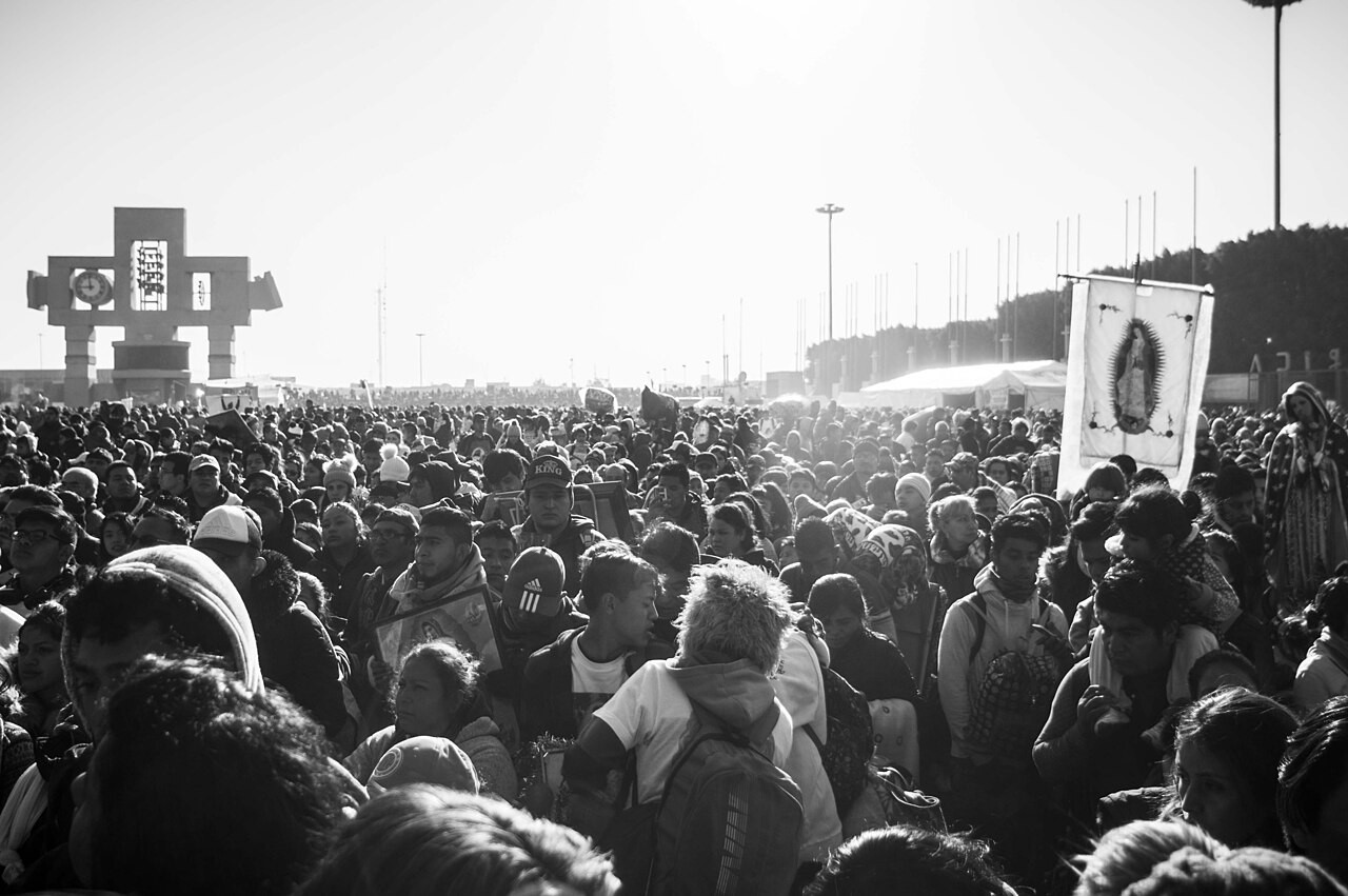 Black-and-white photo of a dense crowd of pilgrims in the plaza of the Basilica of Guadalupe, with a banner of the Virgin and the bell tower behind