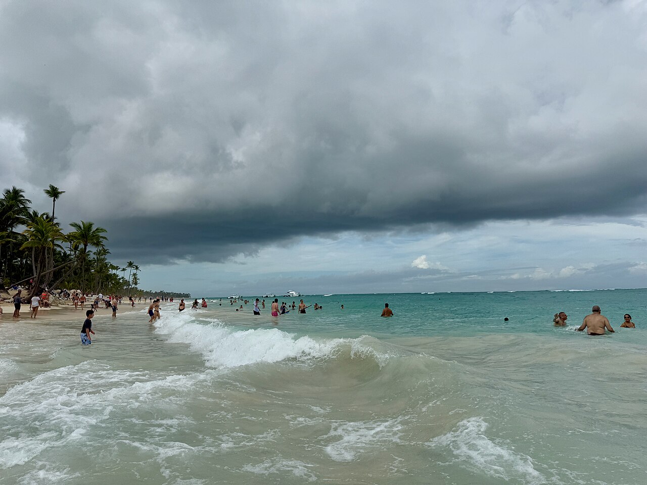 Swimmers in rough green surf under a dark storm cloud on Bávaro beach near Punta Cana, with palm trees along the shore.