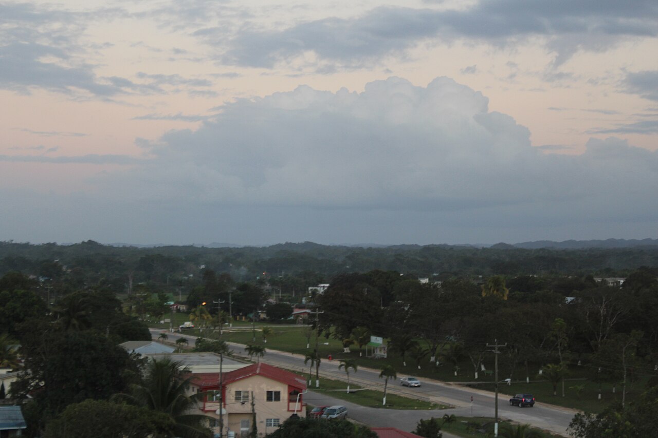 Evening view over Belmopan, Belize's capital, with a road, low houses and palm trees under a large cloud