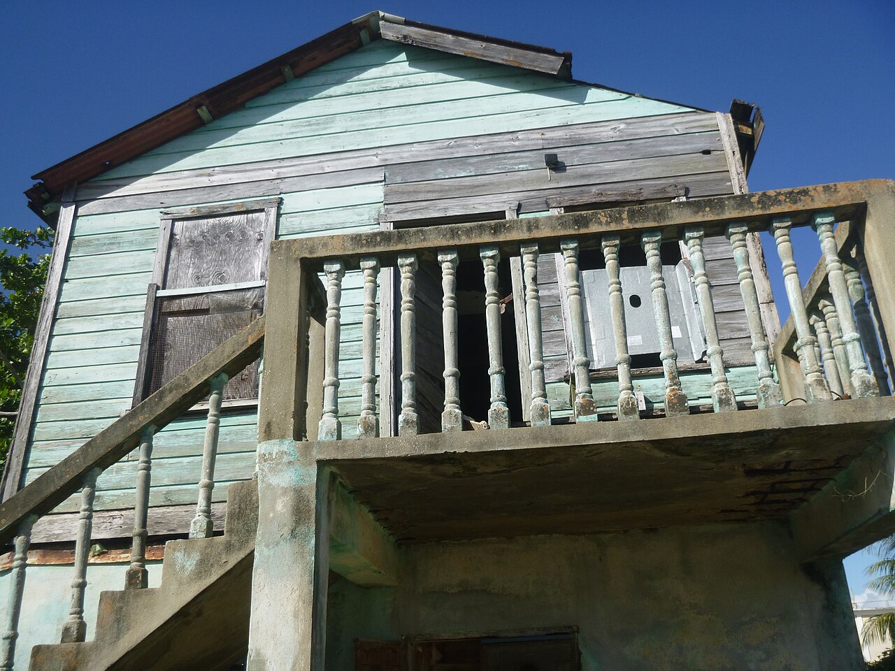 Weathered two-storey wooden house with a concrete balcony and turned balusters under a blue sky in Dangriga, Belize