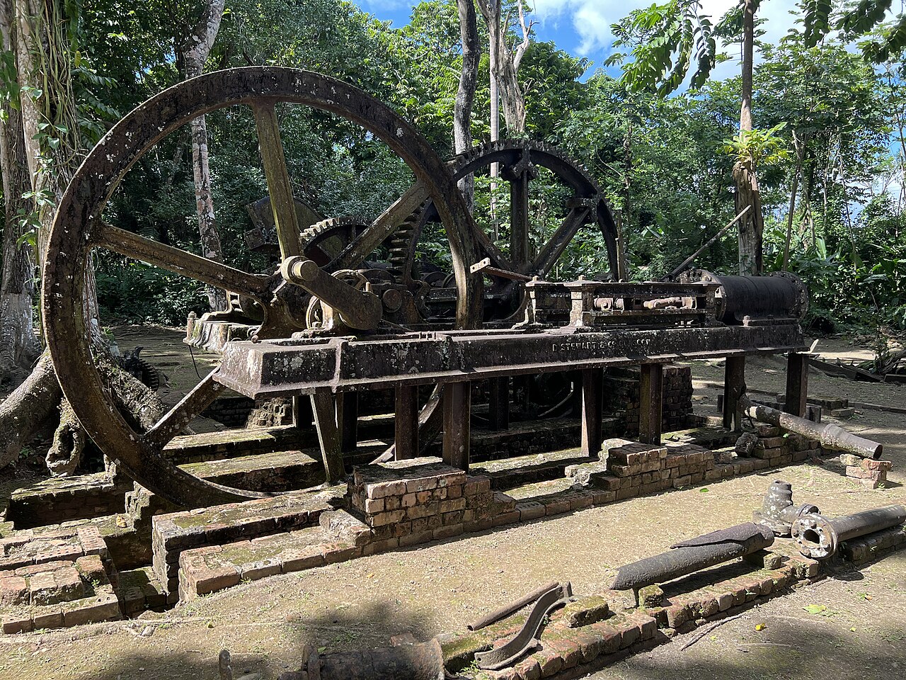 Rusting steam engine and flywheels of the old Serpon sugar mill in Belize, among trees and brick foundations