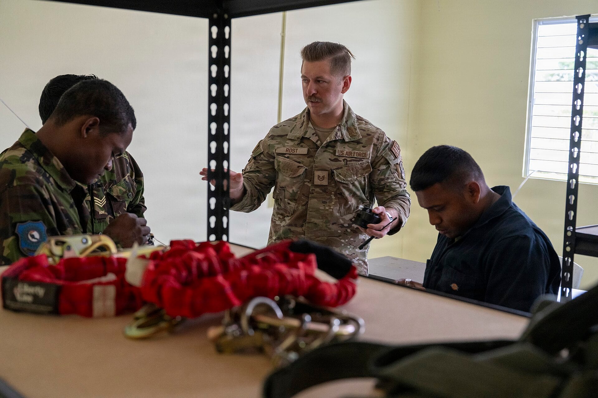 A US Air Force technical sergeant briefing Belize Defence Force members at a table with rigging straps and hooks