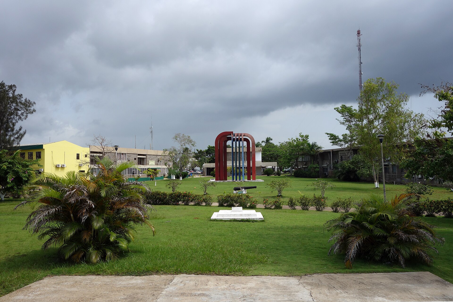 Independence Plaza in Belmopan with a red and blue arched monument on a lawn under storm clouds