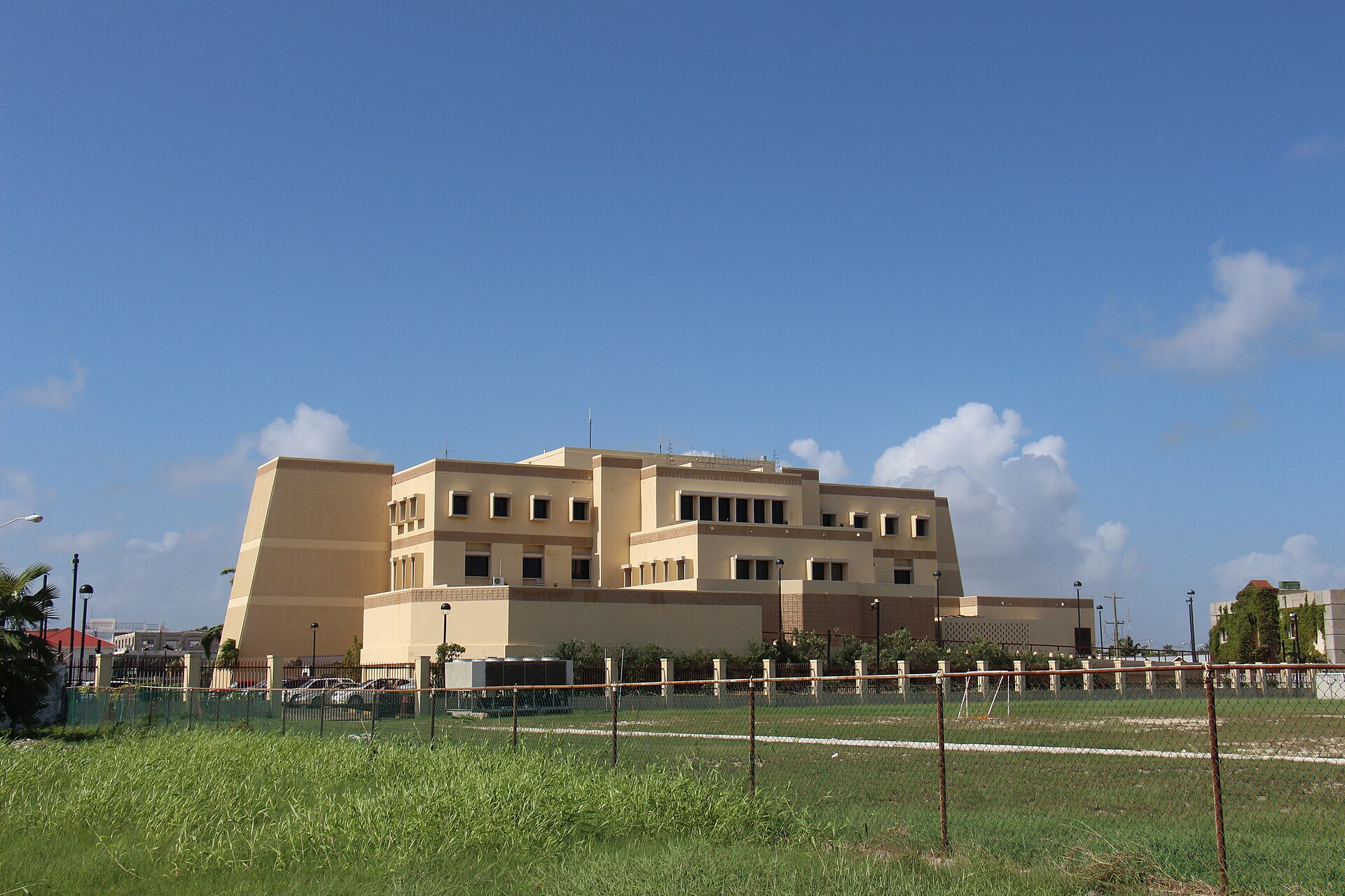 The beige Central Bank of Belize building in Belize City under a blue sky behind a fence and a grass field