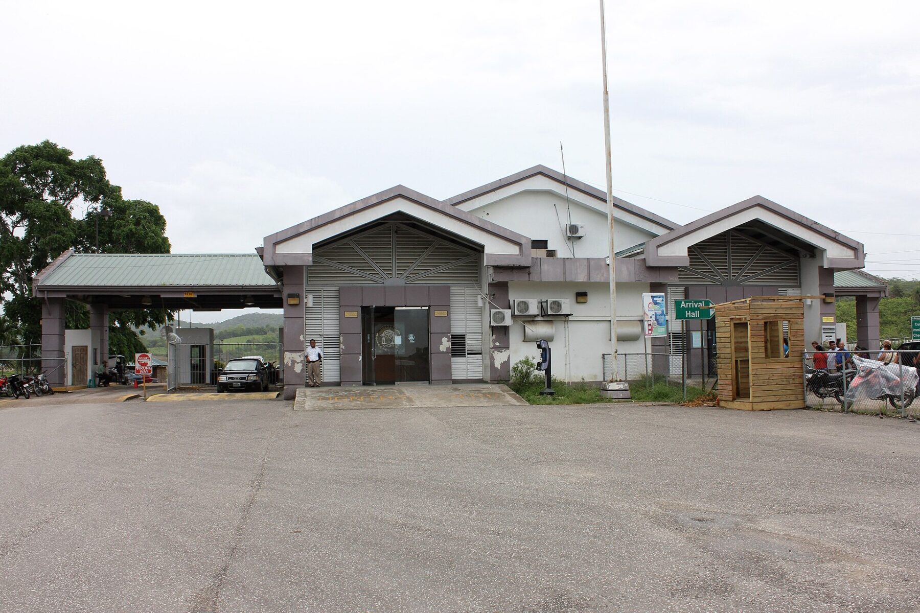 The single-storey Belize border station at Benque Viejo del Carmen, with an Arrival Hall sign, parked cars and wooded hills behind