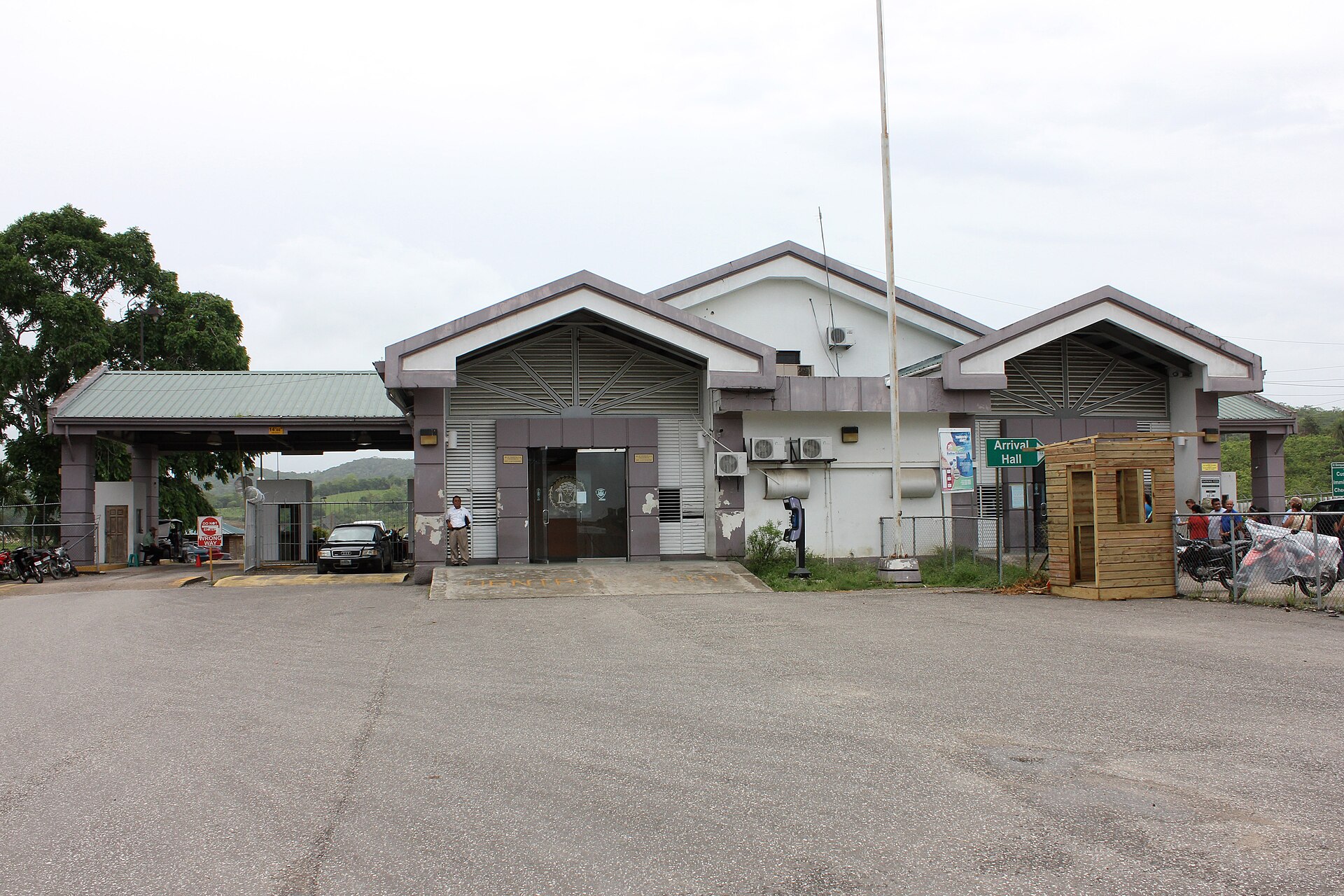 The single-storey Belize border station at Benque Viejo del Carmen, with an Arrival Hall sign, parked cars and wooded hills behind