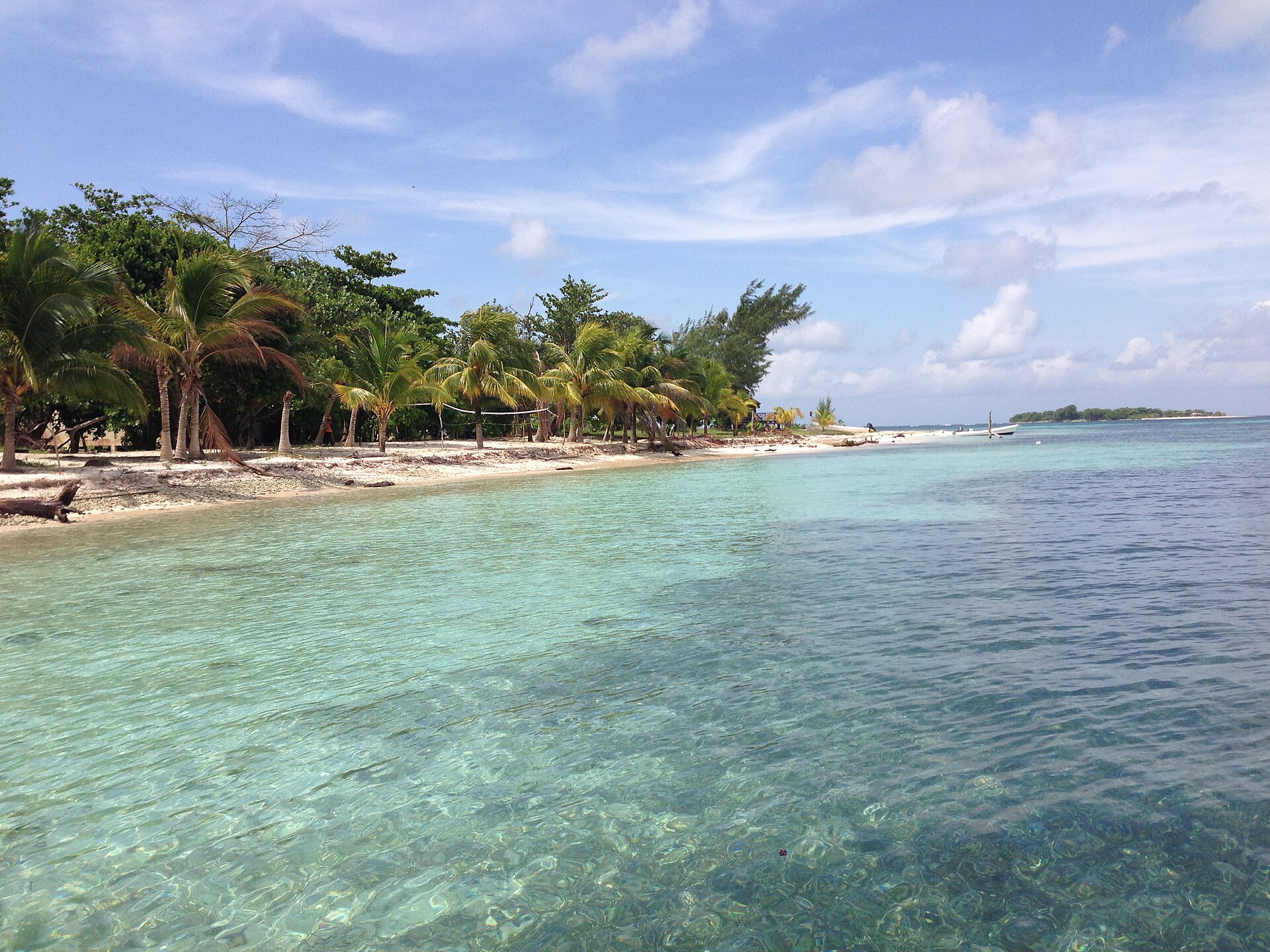 Palm-lined white beach and clear turquoise water on Hunting Caye, with a small boat and another low island on the horizon