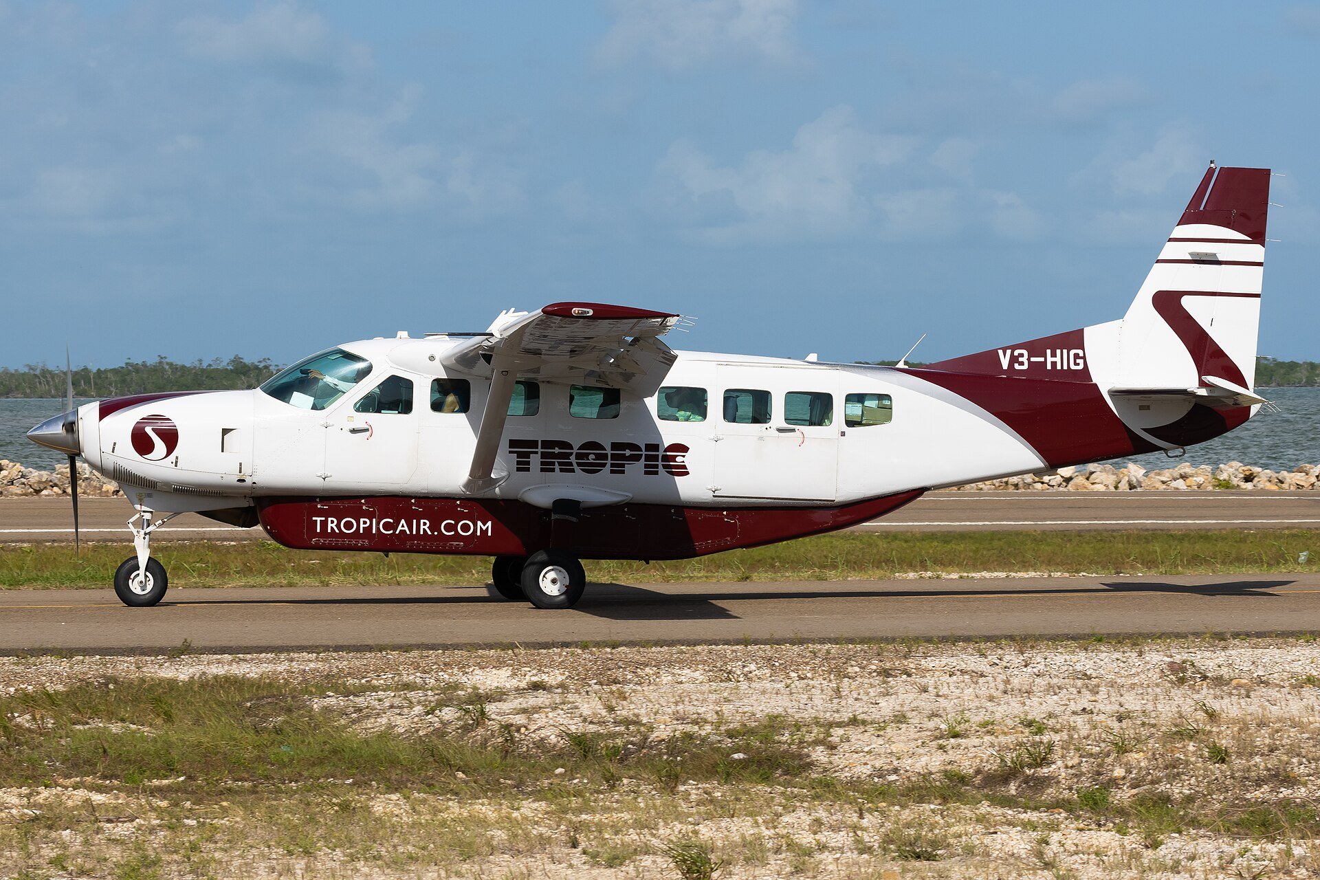 A white and maroon Tropic Air Cessna Grand Caravan aircraft taxiing on a runway beside a lagoon in Belize