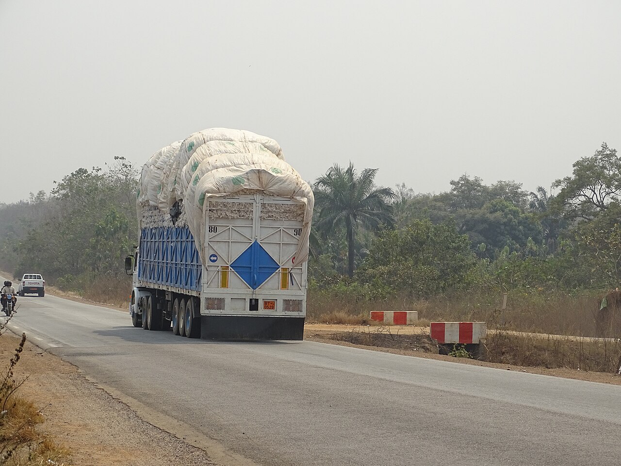 A lorry stacked with white bags of raw cotton driving along a paved road in Benin, with a motorcyclist and a pickup in the distance