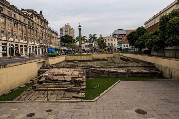 Stone ruins of the Valongo Wharf, a sunken archaeological site in central Rio de Janeiro, with a tall column, old buildings and a green bus on the street behind.