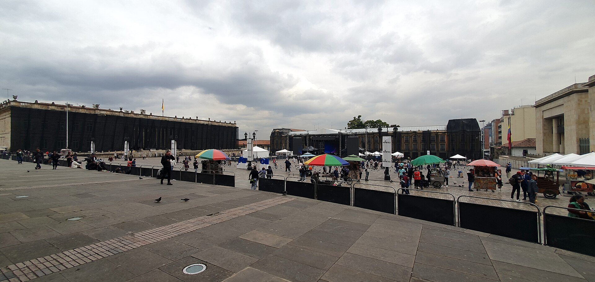 Wide view of the Plaza de Bolívar in central Bogotá with market stalls, a stage and pigeons under a cloudy sky