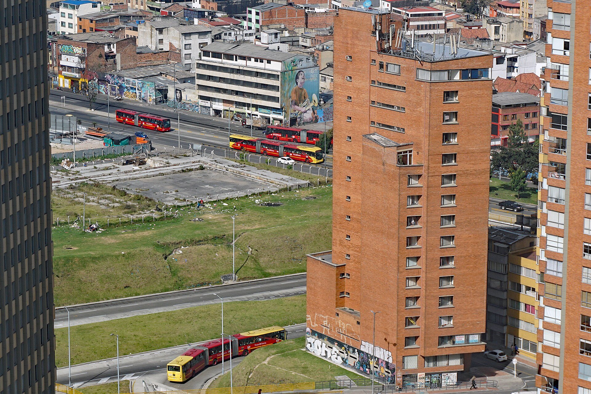Articulated red and yellow TransMilenio buses on Avenida Caracas in Bogotá seen from above, beside brick apartment towers and murals