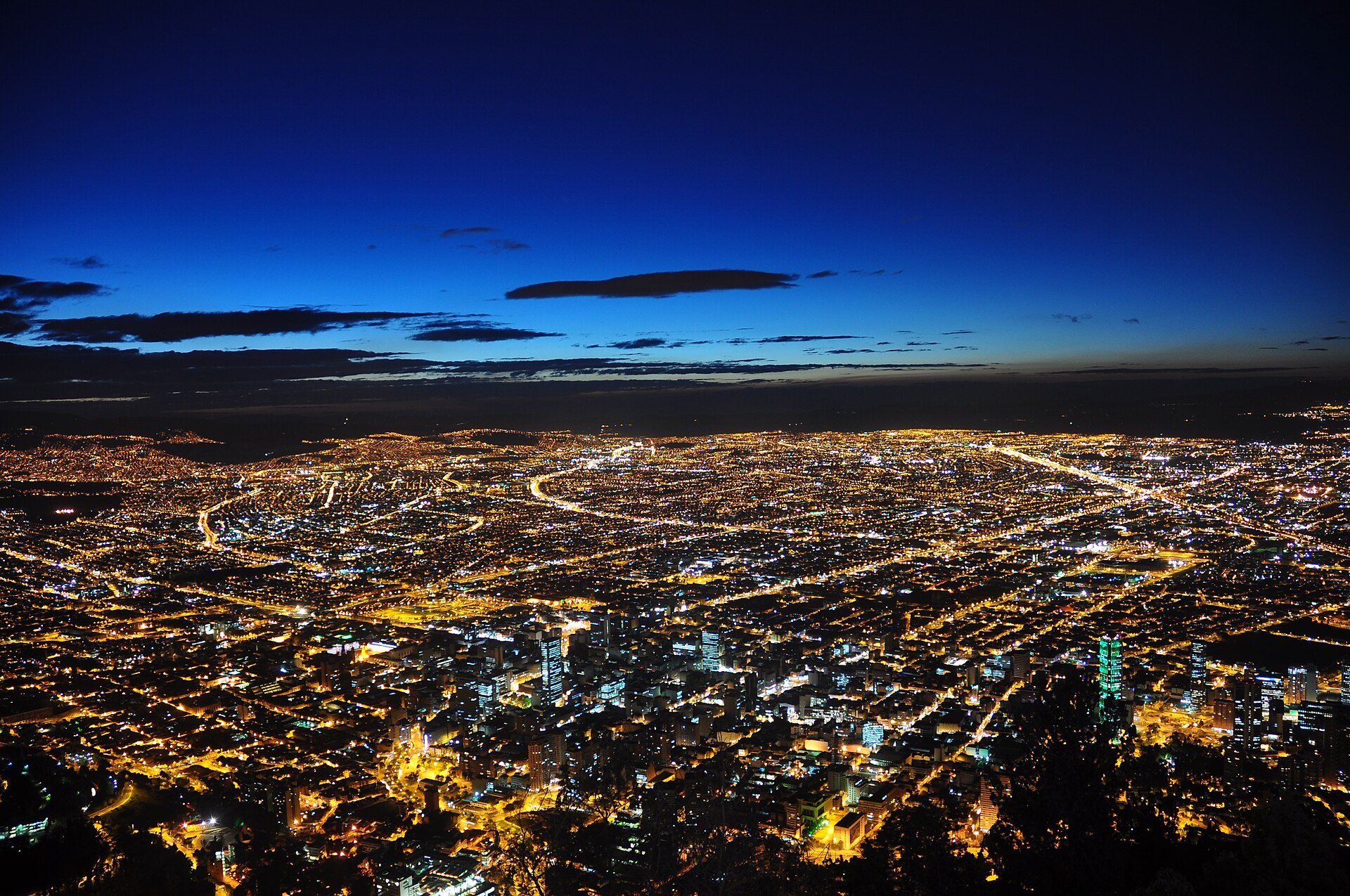 Bogotá at dusk seen from above, with the lights of the city and its office towers spreading toward the horizon under a deep blue sky